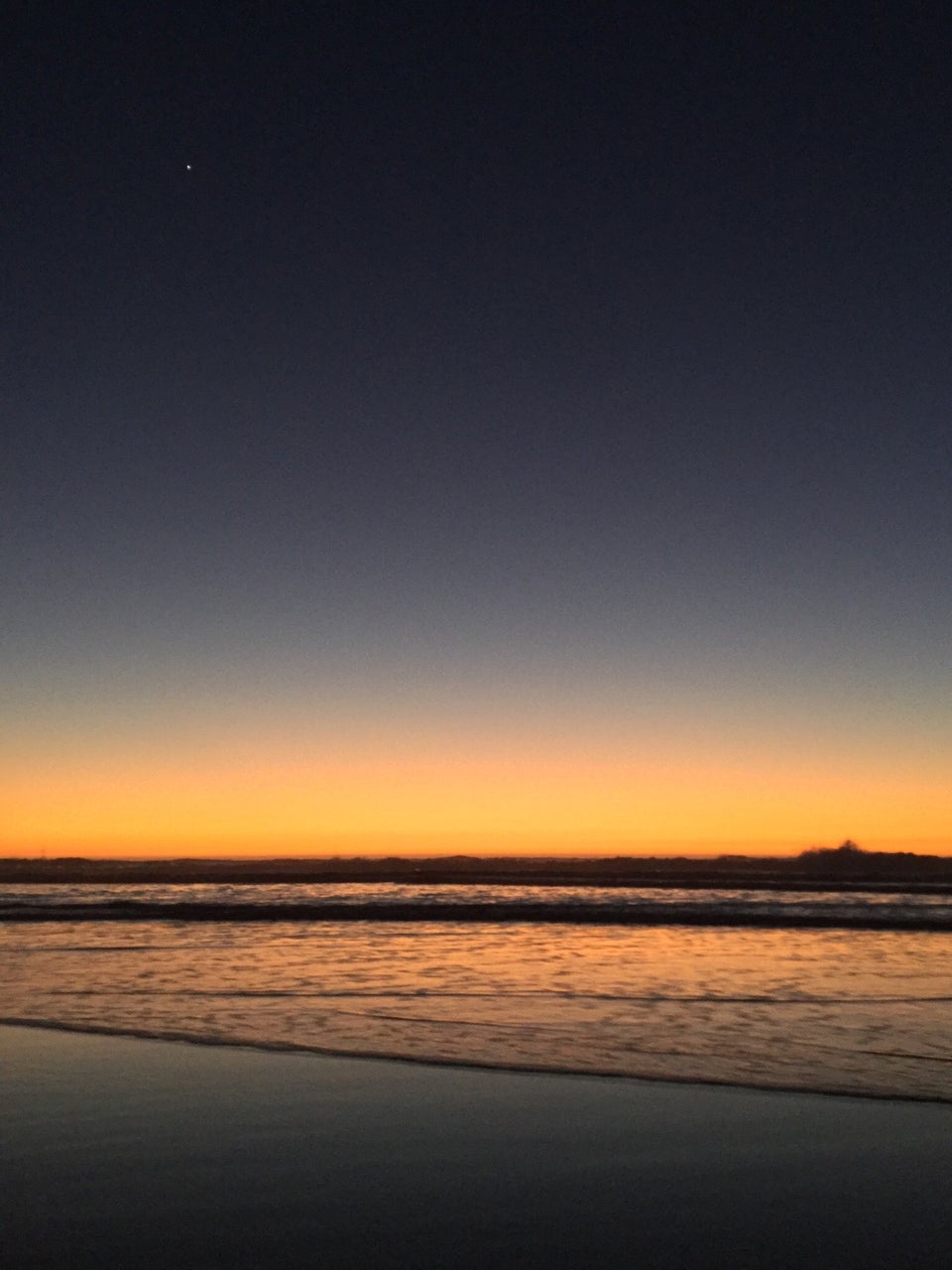 Very dark blue sky with a gradient to a very bright orange at the horizon just above crashing ocean waves, the water also glowing orange, above wet sand reflecting a light to dark gradient.